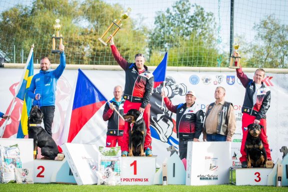 Drei Gewinner auf dem Podest mit Trophäen, umgeben von Flaggen und Hunden.